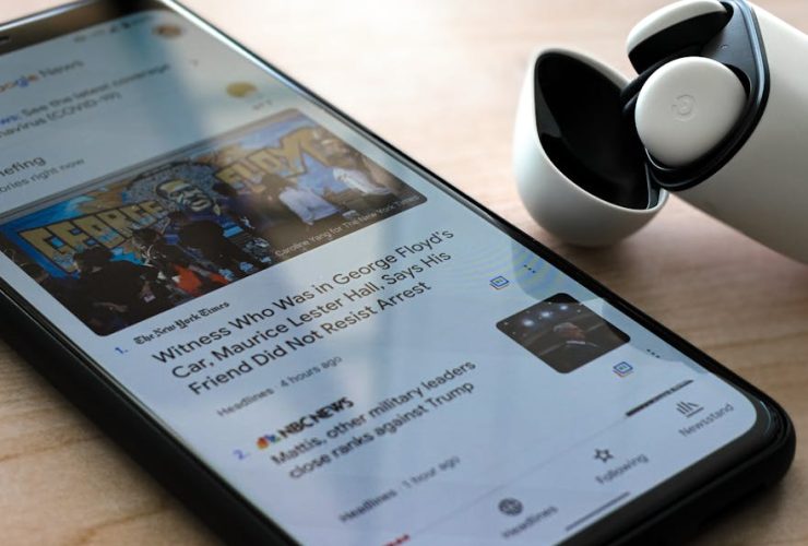Close-up of a smartphone displaying news with wireless earphones on a table.
