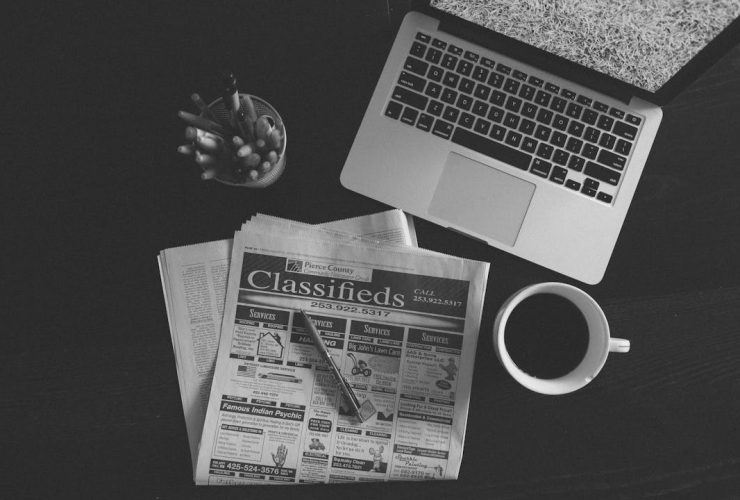 A black and white image of a laptop, newspaper, and coffee cup on a desk.