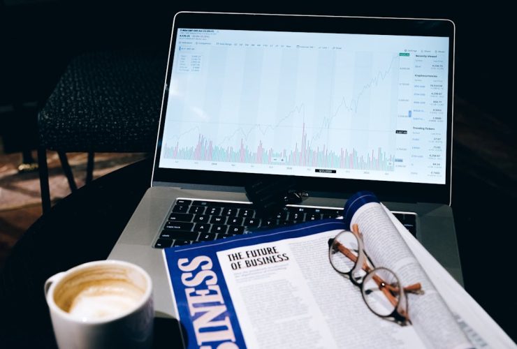 A modern business workspace featuring a laptop showing stock data next to a business magazine and coffee.