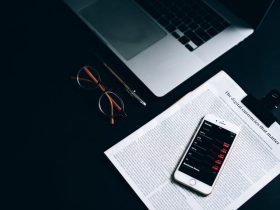 Flat lay of a digital workspace featuring a smartphone, glasses, and a laptop on a dark desk.