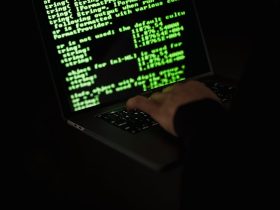 Silhouette of a hacker typing on a laptop in the dark with green code on screen.