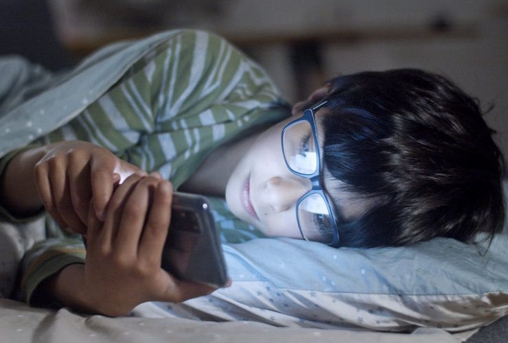 A child lying in bed using a smartphone at night, wearing glasses.