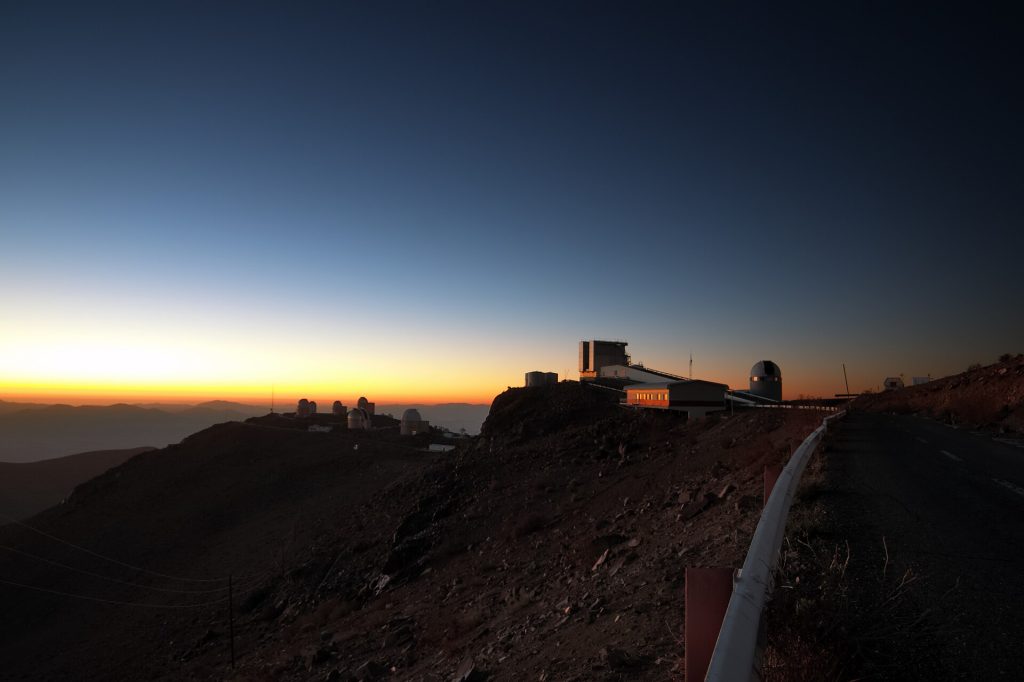 Rays of sunshine illuminate the faces of support buildings and telescopes, including ESO's New Technology Telescope (NTT) dis